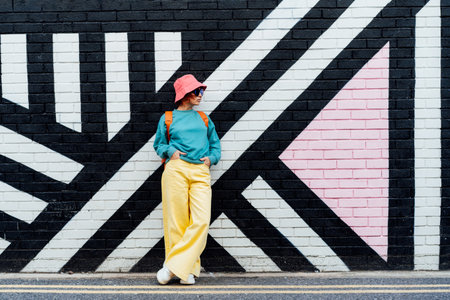 Hipster fashion young woman in bright clothes, sun glasses and bucket hat posing on the painted brick wall background. Urban city street fashion. Selective focus. Copy spaceの写真素材