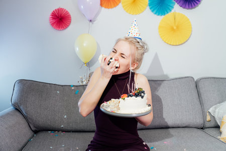 Woman in festive dress and party cap eating birthday cake with her hands, her face stained cream. A...