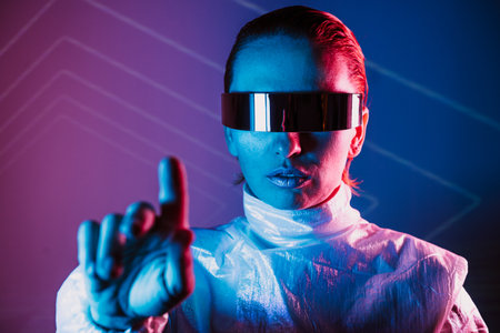 Neon portrait of woman, dressed in futuristic holographic clothes and VR glasses touch space by finger. Cyberpunk girl with virtual touching pad. Neon blue and pink light. Virtual reality glassesの写真素材