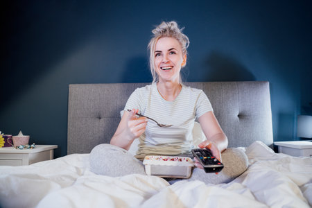 Laughing cheerful woman in home clothes eating ice cream and watching movie on cable TV while...