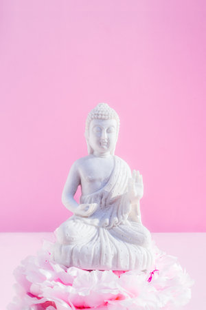 Decorative white Buddha statuette sitting on the peony flower pillow altar on the pink background. Meditation and relaxation ritual. Buddha birthday. Minimalism.Vertical card. Selective focus.の写真素材