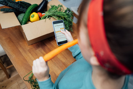 Woman holding phone with active online mobile app of Veganuary diet calendar and eating fresh carrot above box with vegetables. Healthy eating, weight loss. Food delivery, recipe box. Selective focus.の写真素材