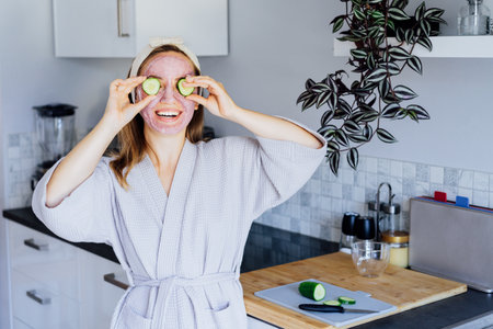 Smiling woman with pink clay facial mask holding just cutted cucumber slices making a refreshing eye mask on the kitchen. Natural cosmetic procedures for skin at home. Funny face. Beauty self-care.の写真素材