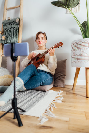 Young Woman playing ukulele guitar, recording music video on phone in modern Scandi interior home. Blogger singing song, content creating. Online learning, teaching, performing, entertaining conceptの写真素材