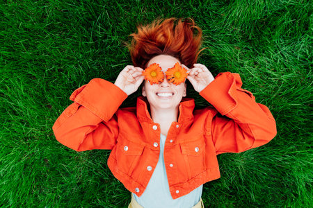 Top view happy smiling redhead Woman with freckles and red orange gerbera Flower glasses lying on green grass. Positive Emotion people. Fashion girl in bright colors jeans clothes. Spring, summer moodの写真素材