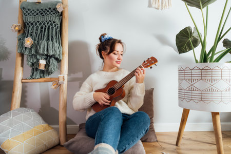 Young woman playing ukulele guitar while relaxing on floor cushion in modern scandinavian home...