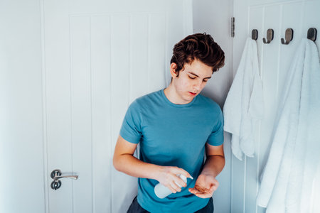 Concentrated caucasian teenage boy with acne problem take care his face skin at home. Cleaning face with washing lotion in bathroom. Teenager skin care process. Selective focus, copy space.の写真素材