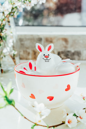 Funny Easter eggs containers with bunny ears filled with candy and chocolate in bowl on white kitchen table with blooming branches. Festive spring Easter composition. Vertical card. Selective focusの写真素材