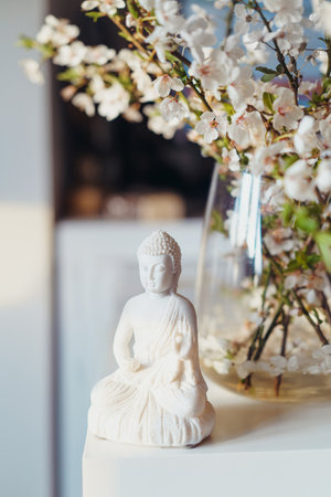 Decorative white Buddha statuette with blooing tree branches in the vase on the background. Meditation and relaxation ritual. Buddha birthday. Vertical card. Selective focus.の写真素材