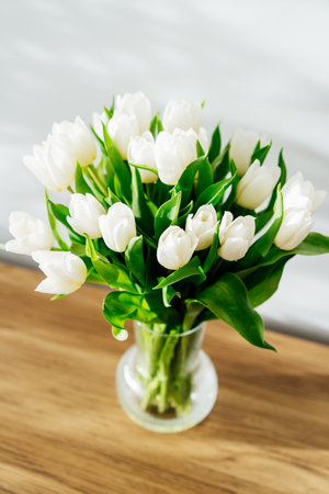 Spring bouquet of white tulip flowers in vase stand on a wooden table near light grey wall with highlights and shadows. Gift for holiday, birthday, 8 March, Mothers Day. Top view. Selective focus.の写真素材