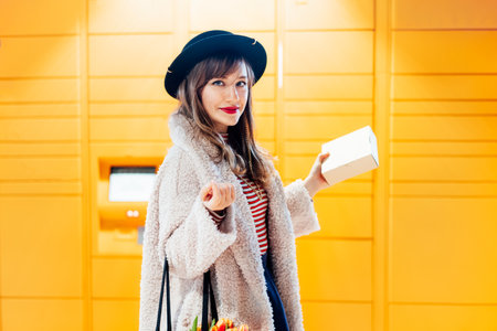 Portrait of smiling fashion woman with box near modern postal automatic mail terminal with self...