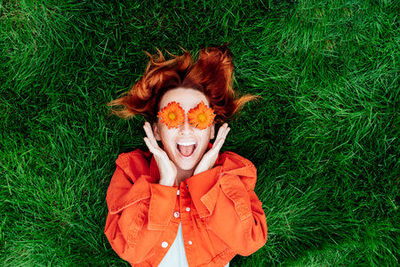 Wow. Emotional surprised, shocked redhead woman with red orange gerbera Flower glasses lying on green grass. Positive Emotion people. Fashion girl in bright colors jeans clothes. Spring, summer moodの写真素材