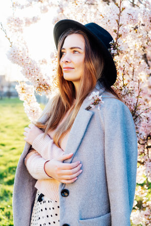 Portrait of a positive, smiling young caucasian woman in hat and coat near a blooming tree with sunset light and sunbeams. Generation Z girl enjoy springtime mood. Selective focus, copy space.の写真素材