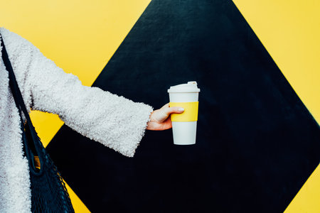 Womans hand holding reusable white coffee cup on the geometry bright yellow and black wall...
