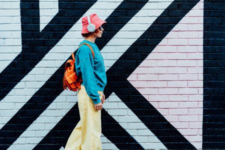 Side view stylish woman in bright clothes and bucket hat wearing wireless headphones and listening to music while walking near city urban painted wall. Fashionable hipster lifestyle. Selective focus.の写真素材