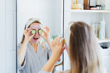 A young smiling woman with pink clay facial mask holds cucumber slices making a refreshing eye mask in bathroom. Natural cosmetic procedures for skin care at home. Beauty self-care. Selective focusの写真素材