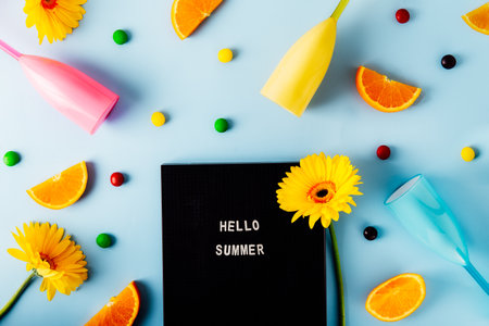Top view flat lay of Letter board with Hello summer inscription, yellow gerbera flower, orange slices, bright cocktail glasses, multicolor candy drops on the light blue background. Summer party mood.の写真素材