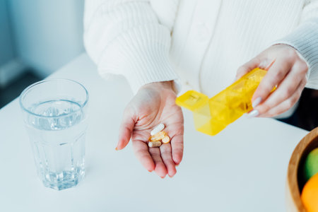 Close-up of womans hand holding pills and pill organizer at home kitchen with water glass. Daily intake of antioxidant diet, vitamin supplements for beauty, skin, hair, healthcare, drugs concept.の写真素材