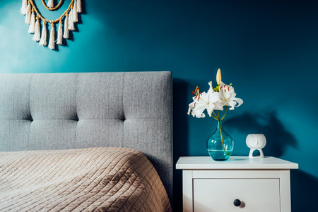 Cozy bedroom with turquoise blue walls and Boho home interior decor: macrame wall hanging decoration, white bedside table,vase with lilly flower, aroma lamp. Dark modern stylish room. Selective focus.の写真素材