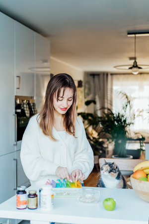 Woman sorting pills into pills organizer with her cat on the kitchen. Pill box with daily doses of medicines. Antioxidant diet vitamin supplements for health and beauty care, biohacking concept.の写真素材