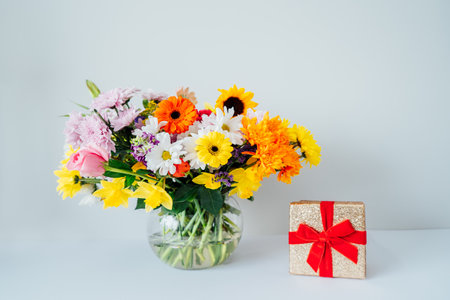 Round Vase with huge multicolor various mixed flower bouquet and shining golden gift box with red velvet ribbon on the white background. Mothers day, women day, valentines day, birthday gift.の写真素材
