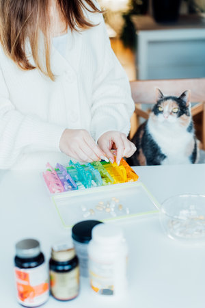 Woman sorting pills into pills organizer with her cat on the kitchen. Pill box with daily doses of medicines. Antioxidant diet vitamin supplements for health and beauty care, biohacking concept.の写真素材