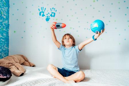 Concentrated boy playing with space rocket and planet Earth models on bed with background of stars and family hand prints on the wall. Concept of education, future, global travel and transportation.の写真素材