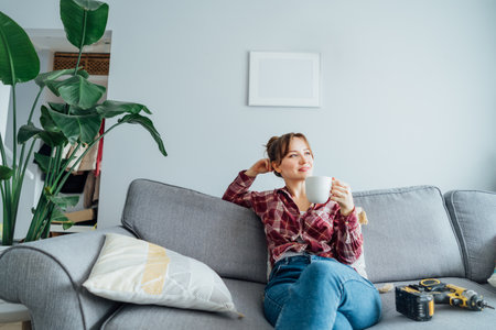 Happy smiling proud of herself young woman relaxing with coffee cup after she puts by her own a picture on the wall at her home. Housekeeping work. Doing repair herself. DIY, gender equality conceptの写真素材