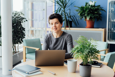 Neutral gender middle aged woman with no make-up in casual clothing using laptop and smiling while working indoors in her workstation in an open space office. Online video call, IT HR, recruitingの写真素材