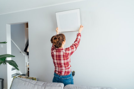 Back view young woman putting painting, mockup picture frame on the wall. Casual lady taking care of coziness in her new stylish apartment. Young female decorating room. Moving and renovate houseの写真素材