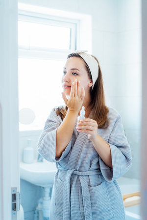 Young woman in bathrobe looking in the mirror, holding bottle with hyaluronic acid serum and applying it on face. Skin hydrating. Cosmetic spa procedures. Beauty self-care at home. Selective focusの写真素材