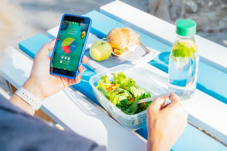 Close up woman using meal tracker app on phone while eating salad at picnic table in the park on a break. Healthy balanced diet lunch box. Healthy diet plan for weight loss. Selective focusの写真素材