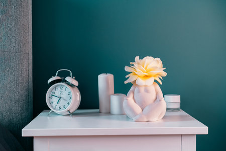 Closeup white bedside table with clock, female body-shaped vase with rose flower and candles on turquoise wall background. Modern stylish female bedroom. Self recovery place at home. Selective focus.の写真素材