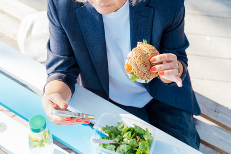 Cropped business woman eating wholemeal burger with salad and using a phone sitting at picnic table in the park while having a break. Healthy lunch box for weight loss. Balanced diet. Selective focus.の写真素材