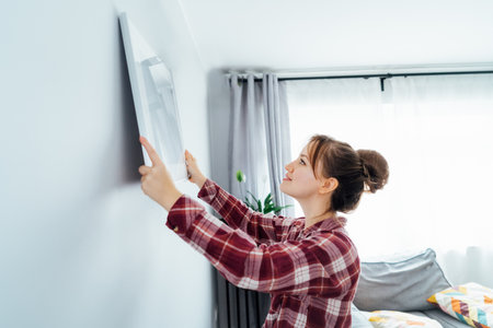 Side view young woman putting painting, mockup picture frame on the wall. Casual lady taking care of...