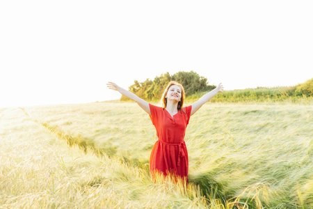 A happy young woman with raised arms relaxing in wheat field on sunset. Celebrating freedom. Positive emotions feeling life, peace of mind. Mental health practice. Nature relaxation. Selective focus.の写真素材