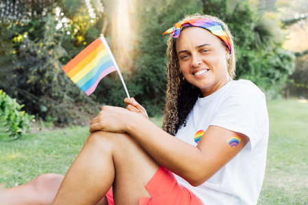 Mixed race woman with small rainbow flag outdoor. Young lesbian activist smiling and holding flag symbol of social movement Lgbtq. Fight for equality, freedom and human rights. Celebrating Pride monthの写真素材