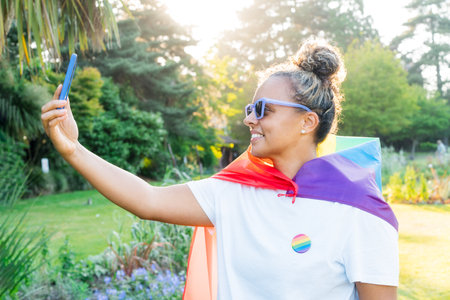 Young Latin happy woman wrapped in a rainbow flag, symbol of LGBT community, makes selfie on a...