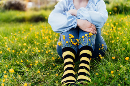 Close up young female wearing jeans and striped black and yellow socks with flowers inside sitting on the green grass of blooming meadow. Concept of bee protection, bloom season, art, creativityの写真素材