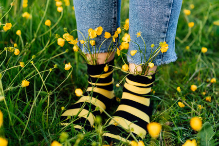 Close up female feet wearing jeans and striped black and yellow socks with flowers inside standing on the green grass of blooming meadow. Concept of bee protection, bloom season, art, creativity.の写真素材