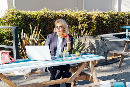 Middle-aged business woman having lunch at the park outdoor cafe, picnic area and working on laptop during her break. Balanced diet lunch box. Healthy eating habits and well-being. Selective focusの写真素材