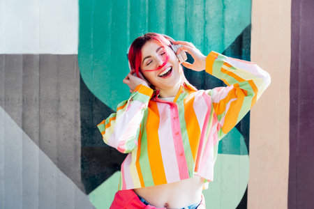 Emotional stylish young woman with pink hair in multicolor striped shirt wears wireless headphones, listening to music and singing on a graffiti wall background. enjoy the moment. Fashionable hipsterの写真素材