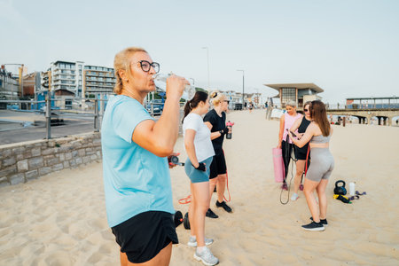 Middle aged woman drinking water from reusable glass bottle while taking rest, refreshing during group workout class exercise with coach on the beach. Sport for health and wellbeing. Active lifestyleの写真素材