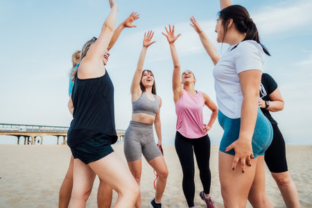 Happy fitness class giving high five after completing workout on beach. Multi aged women motivated after session together, engaged in team building, join hands for shared goal or success at trainingの写真素材