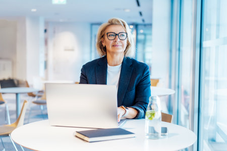 50s confident mature businesswoman dreamy looking at window, middle-aged experienced senior female professional working on laptop in open space office. Female entrepreneur working remotelyの写真素材