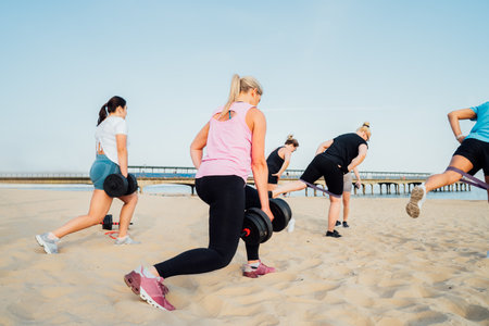 Back view women of various ages doing fitness workouts in class exercise with coach on the beach. Ladies working with dumbbells and resistance bands. Sport for health and wellbeing. Active lifestyle.の写真素材
