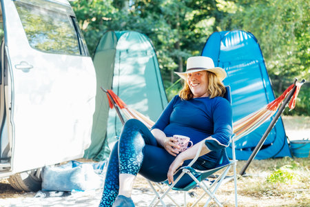 Adult woman relaxing, drinking tea while sitting in the travel chair near camper car vehicle and tents. Weekend trip activity. Female living on camper car and travel the world. Caravan car Vacationの写真素材