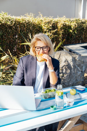 Middle-aged businesswoman having lunch at the park outdoor cafe, picnic area and working on laptop during her break. Balanced healthy diet lunch box. Unhealthy eating habits. Bad work-life balance.の写真素材