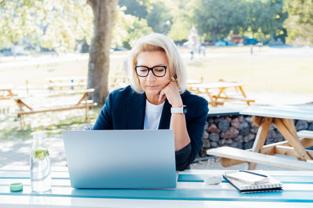 Concentrated 50s stylish, confident mature business woman, middle aged lady using wireless ear buds and laptop, working remotely in park. Caucasian woman make a video call, study, work online lesson.の写真素材