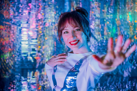 Happy smiling woman in white clothes dancing in neon light and extends a hand inviting you. Music lover. White dress code party, disco. Enjoy moment on shiny background. Selective focusの写真素材